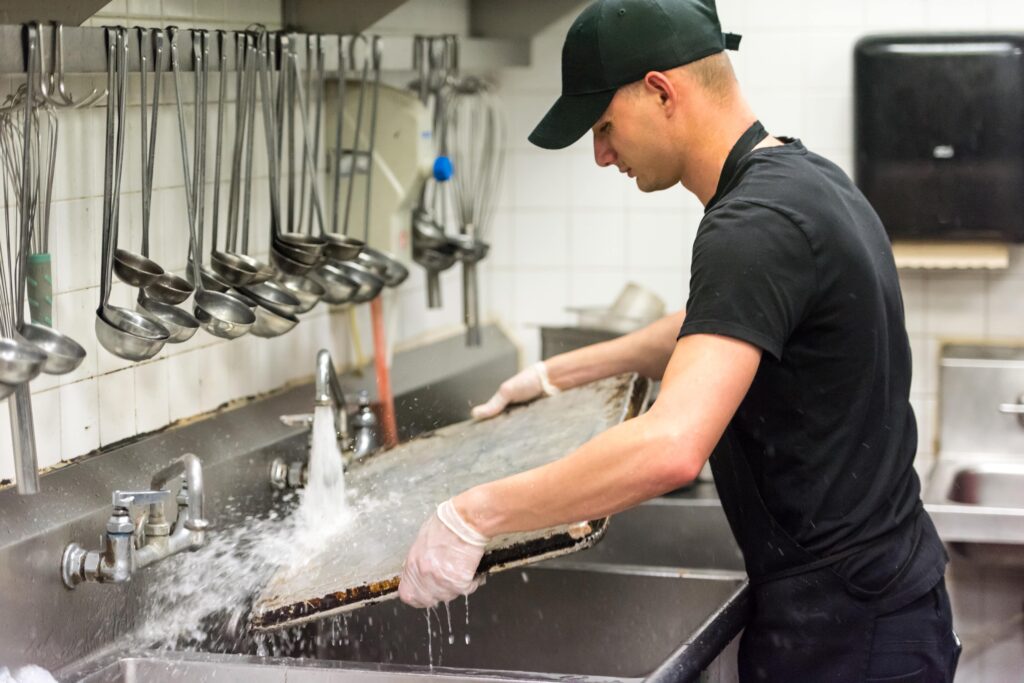 Kitchen worker washing a large baking tray under running water in a commercial sink.