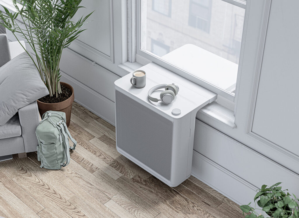 Modern room heat pump unit installed below a window with headphones and a mug resting on top.