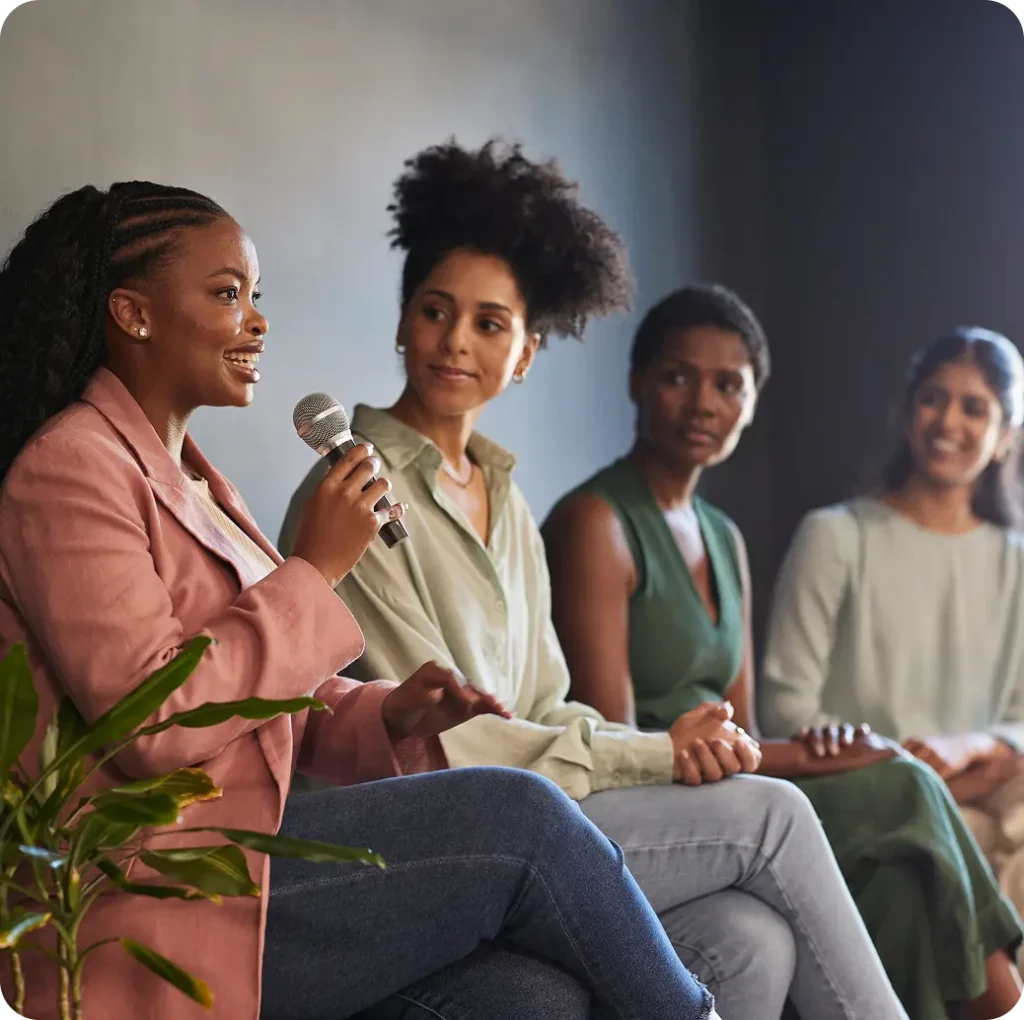 Four women seated on stage, one speaking into a microphone while the others listen attentively
