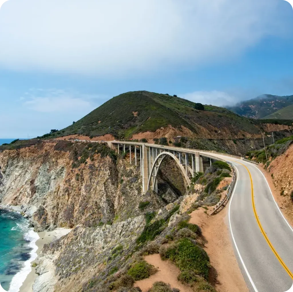 Coastal bridge curving along a cliffside road with the ocean below and green hills in the background in California.