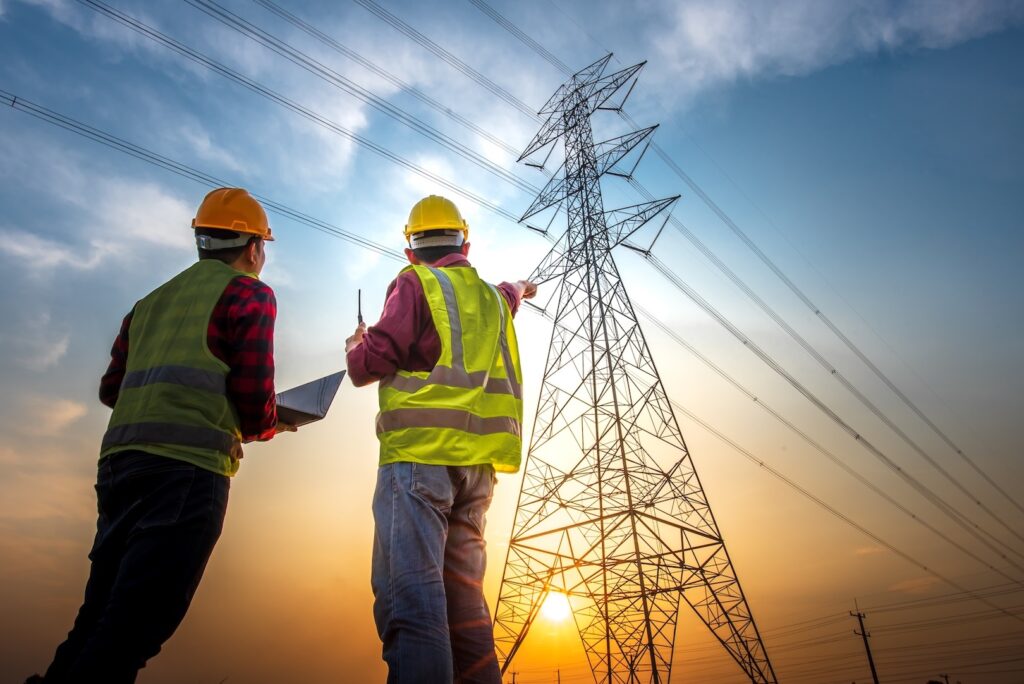Two utility workers inspecting a high-voltage transmission tower at sunset.